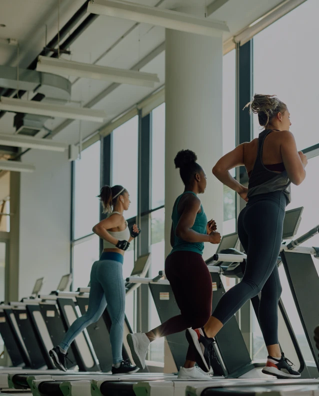 Three women running on treadmills in a bright, modern gym with large windows