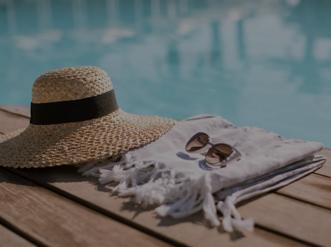 Straw sunhat, white towel, and sunglasses resting poolside on a wooden deck