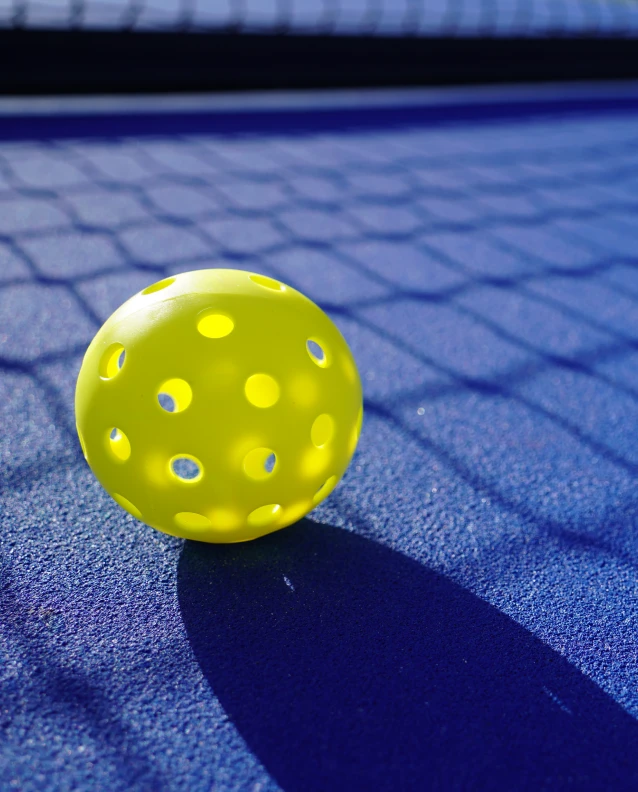 Bright yellow perforated pickleball resting on a vibrant blue court under strong sunlight