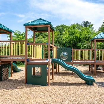Large kids’ playground with green slides, climbing features, and mulch flooring under sunny skies