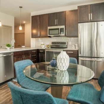 Glass dining table with teal chairs centered in kitchen featuring stainless appliances and pendant lights