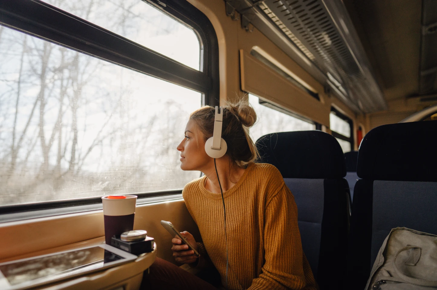 Woman wearing headphones gazing out train window, holding smartphone during travel